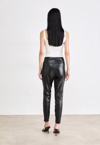 Woman with long black hair wearing a sleeveless white top, black leather pants, and black high-heeled sandals, standing against a plain background.