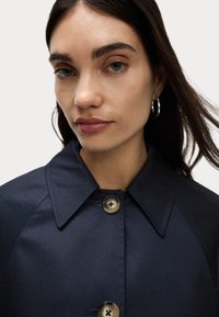 Cappotto leggero di colore blu navy, con colletto a punta e due grandi bottoni. Tessuto liscio, cuciture dettagliate sulle spalle.