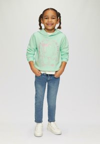 Jasnozielona bluza z kapturem z różowym wzorem graficznym, noszona na białej koszuli, w połączeniu z niebieskimi dżinsami i białymi trampkami.