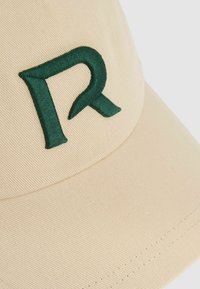 Beige baseball cap with a structured crown and curved visor. Features green embroidered letter "R" on the front. Textured fabric material.