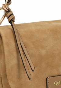 Tasche aus tanfarbenem Wildleder mit glatter Textur, ausgestattet mit einem goldenen Logoschild, eleganten Beschlägen und einem abnehmbaren Tragegurt mit zwei langen Quasten.