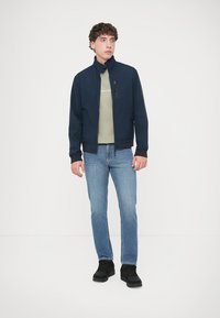 Μπλε ναυτικό μπουφάν μαλακού υφάσματος με ψηλό γιακά και τσέπη με φερμουάρ, συνδυασμένο με ανοιχτό πράσινο πουκάμισο, ξεθωριασμένο μπλε τζιν και μαύρα παπούτσια.