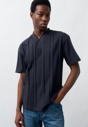 Νεαρός άνδρας που φοράει σκούρο μπλε μπλουζάκι με κοντά μανίκια και κάθετες πιέτες, καθώς και μπλε τζιν, στέκεται μπροστά σε απλό ανοιχτό φόντο.