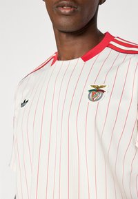 Bílý dres s červenými svislými pruhy, červeným límcem a třemi červenými pruhy na rameni. Na hrudi je vyšitý klubový znak.