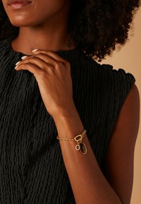 Gouden kettingarmband met een rode edelsteen en een ronde bedel, tentoongesteld op een gestructureerde zwarte stoffen achtergrond.
