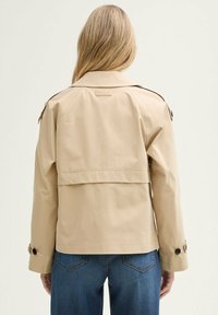 Beige Trenchcoat mit cropped Design, ausgestattet mit einer Rückenschleife, knöpfbaren Ärmeln und einem Etikett am Hals, kombiniert mit blauen Jeans.