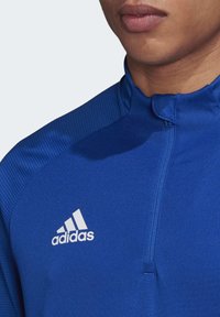 Blå atletisk zip-up top med tekstureret stof på skuldrene, slankt design og hvid Adidas-logo på venstre side.