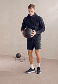 Una palla medica nera pesante tenuta da una persona in una felpa nera con zip e pantaloni corti neri. Il pavimento è neutro, con un manubrio e una piccola palla nelle vicinanze.