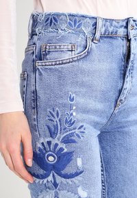 Světle modré džíny z denimu s třepeným pasem a složitou modrou květinovou výšivkou na levém stehně. Pět kapsový design, pohodlný střih.