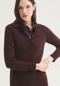 Jeune femme aux cheveux bruns et lisses portant un pull marron foncé à manches longues par-dessus une chemise bleue à motifs avec col, debout avec une main sur la taille.