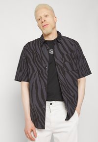 Black and gray short-sleeve button-up shirt with a tiger stripe pattern, front pocket, worn over a black t-shirt, paired with white pants.
