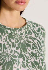 Frau, die einen grün-weißen, floral gemusterten Strickpullover trägt, wobei der Ausschnitt sowie ein Teil des Gesichts und des Nackens zu sehen sind.