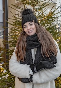 Berretto di lana nera con pompon, sciarpa nera a trama, e cappotto bianco soffice; mani coperte con guanti neri. Albero verde con luci sullo sfondo.