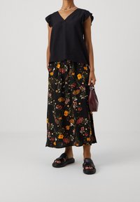 Top nero a maniche corte con scollatura a V, abbinato a una gonna maxi con motivo floreale che presenta colori vivaci su uno sfondo scuro. Sandali neri e una borsa piccola.