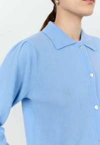 Svetlo modri kardigan, izdelan iz mehkega materiala, z klasičnim ovratnikom, zapenjanjem na gumbe in rahlo napihnjenimi rokavi. Teksturirana površina.