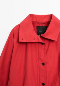 Veste rouge avec un grand col, dotée d'une fermeture éclair à l'avant, de boutons noirs et d'une étiquette indiquant "Massimo Dutti". Texture lisse et matériau léger.