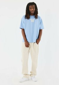 Camiseta de gran tamaño en azul claro con un gráfico sutil, combinada con pantalones beige con detalles de flecos y zapatillas blancas.