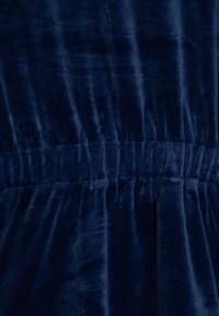 Donkerblauwe fluwelen stof met een gladde textuur, met ruchingdetails bij de taille. Zachte glans met subtiele lichtvariaties.