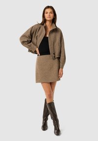 Brun houndstooth-mønsteret jakke med høj krave, parret med en matchende nederdel. Modellen har en sort top og høje brune støvler på.