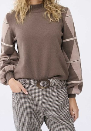 Femme portant un pull marron avec des manches ballon à motifs géométriques et un pantalon pied-de-poule avec ceinture à boucle ovale, main dans la poche.