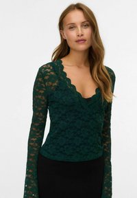 Haut en dentelle verte à manches longues avec un décolleté festonné et un motif floral. Porté sur une couche noire en dessous, le matériau texturé ajoute de la profondeur.