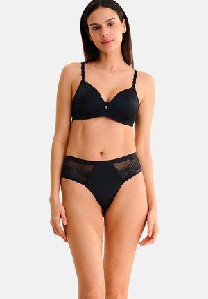 Femme portant un soutien-gorge noir avec fines bretelles en dentelle et une culotte en dentelle noire semi-transparente assortie, debout devant un fond clair uni.