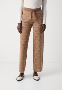 Pantalons larges à motifs marron avec un design texturé, une taille élastique et un détail à nouer. Associés à des chaussures crème à enfiler.