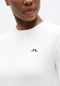 Λευκό εφαρμοστό τοπ με στρογγυλή λαιμόκοψη. Διαθέτει μικρό μαύρο λογότυπο στο στήθος και απαλή, ελαστική υφή υφάσματος.