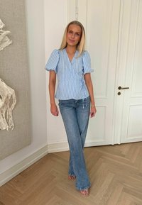 Jeune femme debout à l'intérieur portant un haut cache-cœur rayé bleu avec des manches bouffantes et un jean bleu à jambes larges sur un sol en bois.
