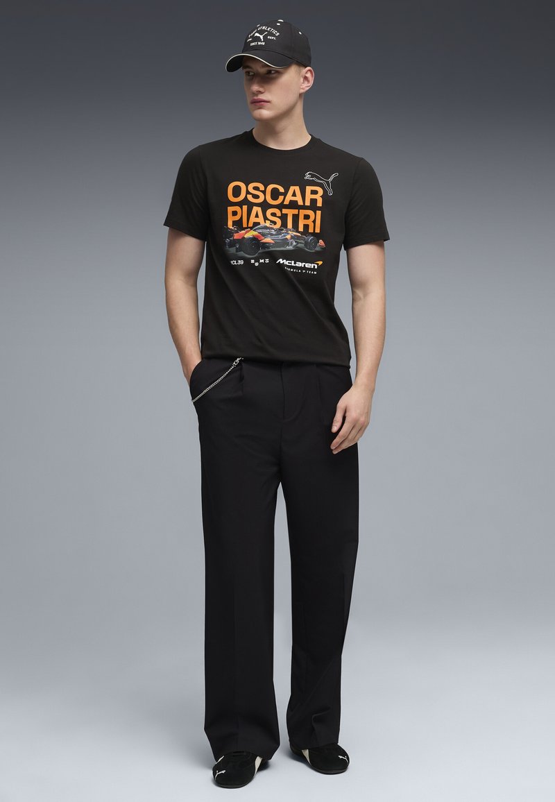 Noor mees, kandes musta Oscar Piastri McLareni T-särki, musti laia lõikega pükse kettaga, musti Puma tossusid ja musta mütsi.