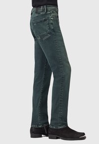 Osoba oblékající tmavě zelené džíny rovného střihu a černé kotníkové boty, stojící před jednoduchým světlým pozadím.