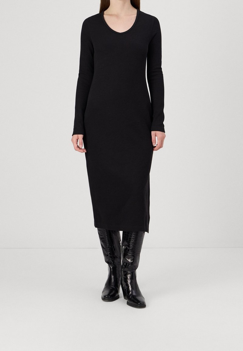 Femme portant une robe midi noire à manches longues, associée à des bottes hautes noires brillantes, debout devant un fond clair uni.