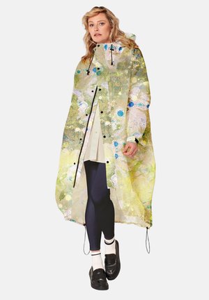 Vrouw draagt een doorschijnende regenjas met een abstract groen, geel en blauw patroon, zwarte legging, witte sokken en zwarte loafers.