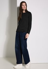 Musta pitkähiha-paita, jossa on tekstuuripinta, yhdistettynä leveälahkeisiin tummiin farkkuihin ja valkoisiin sneakereihin, seisoo vaaleaa taustaa vasten.