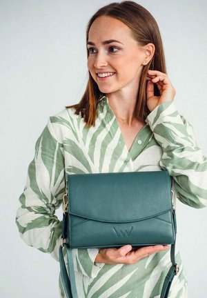 Frau in grün-weiß gemustertem Hemd lächelnd, hält eine tealfarbene Ledertasche mit Schulterriemen vor schlichtem Hintergrund.