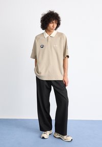 Beige polo shirt with a white collar, loose fit, featuring a circular logo and "1" detail. Paired with black wide-leg trousers and beige sneakers.