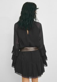 Rochie din voal negru cu mâneci volan, detaliu cu decolteu în formă de cheie la spate și o curea marron cu studuri. Rochia are un tiv fantezist în straturi.