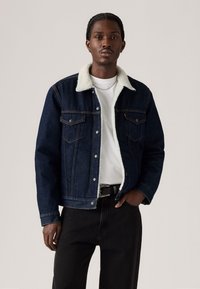 Chaqueta de denim oscuro con un collar blanco y esponjoso; dos bolsillos en el pecho; cierre de botones en la parte delantera; usada sobre una camiseta blanca y jeans negros.