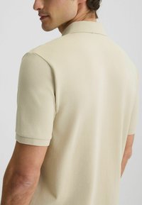 Bež polo majica kratkih rukava, mekane teksture i s ovratnikom. Ima klasičan kroj i minimalistički dizajn.