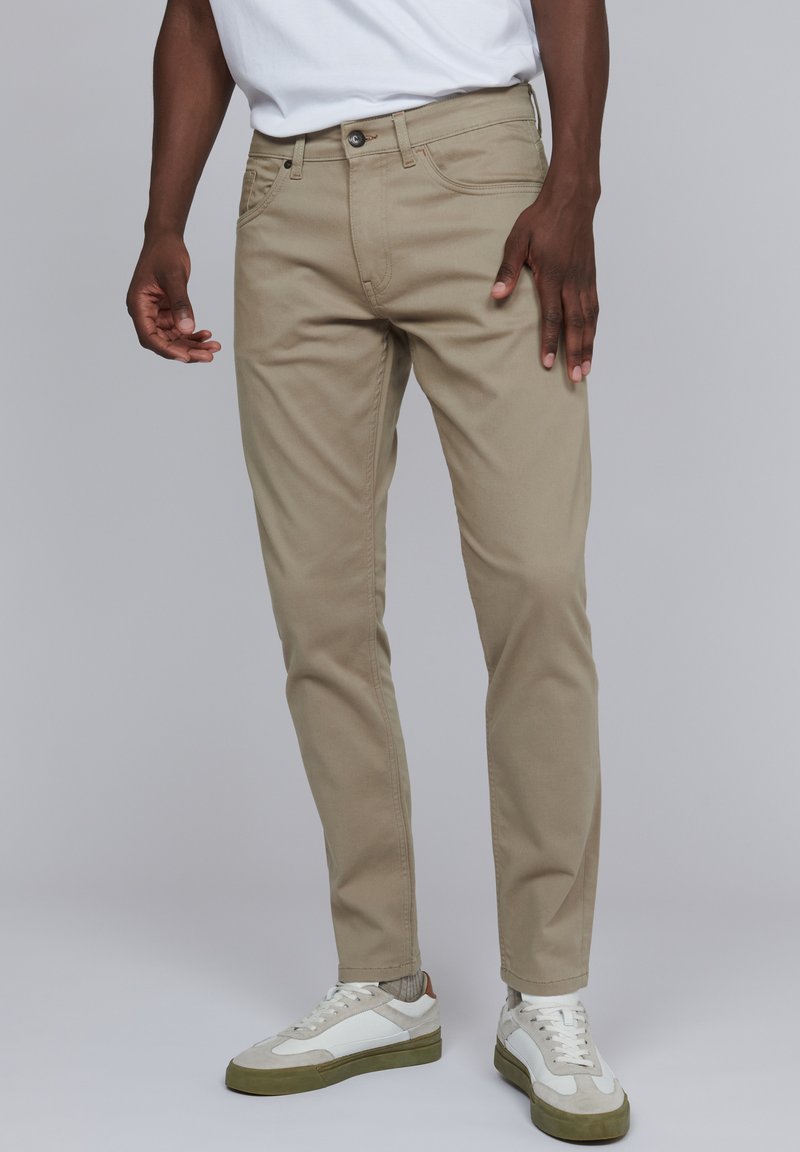 Pantalons en coton beige avec une coupe slim, dotés de poches avant et de passants de ceinture. Associés à des baskets blanches et beiges.