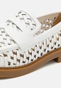 Mocassin tissé en cuir blanc avec semelle beige et détails de couture décoratifs sur un fond clair.