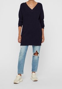 Merinovärinen neulepusero, jossa on V-kaulus ja kolmanneksen pituiset hihat, yhdistettynä vaaleisiin kuluneisiin farkkuihin ja beigeihin lenkkareihin.