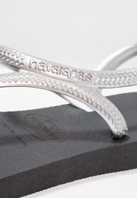 Hopeakylkiset flip-flopit, joissa on kudottu hihnasuunnittelu ja "havaianas"-logo; musta teksturoitu pohja, jossa on ympyräkuvioita ja tuotteelle tyypillisiä yksityiskohtia.