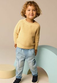 Ljusgul stickad tröja med ribbat mönster, kombinerad med ljusblå denimbyxor med sidfickor och svarta sneakers.
