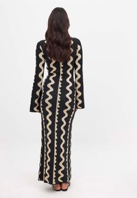 Robe noire à manches longues avec un motif en zigzag crème. Silhouette ajustée, longueur au sol, texture côtelée, et manches évasées. Pas de matériel visible.