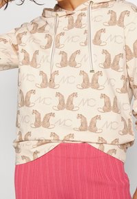 Felpa beige con un motivo a stampa leopardata ripetuto e testo "MC" in marrone chiaro. I lacci e l'orlo a coste aggiungono dettagli.