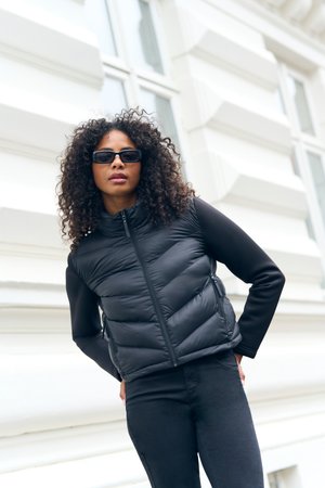 Femme aux cheveux bouclés portant des lunettes de soleil noires, une doudoune noire et un pantalon noir, debout à l'extérieur contre un mur blanc d'un bâtiment.