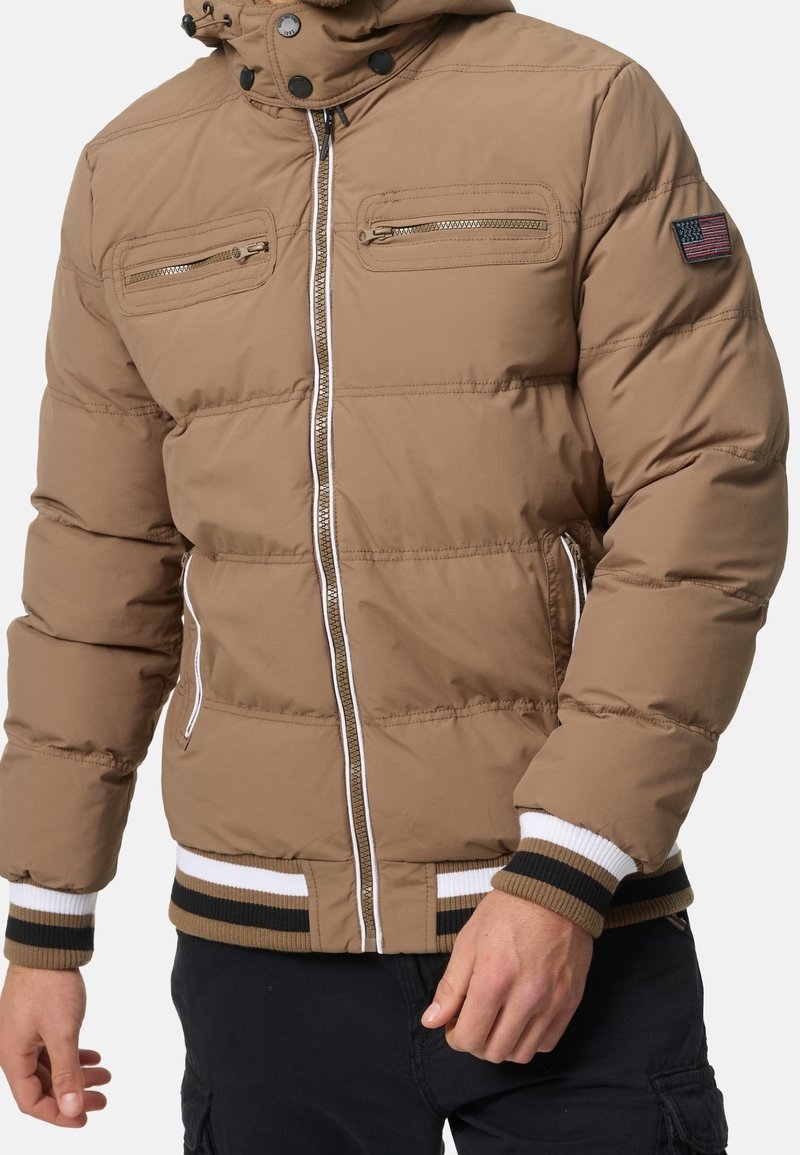 Beige gepolsterte Jacke mit Frontreißverschluss, zwei mit Reißverschluss versehenen Brusttaschen, gestreiften ribbeligen Bündchen und Saum sowie einem Aufnäher mit der amerikanischen Flagge auf dem linken Ärmel.