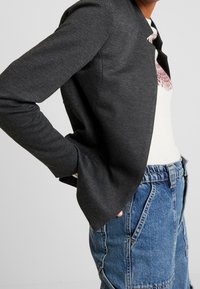 Blazer gris foncé avec un motif texturé, présentant une coupe slim et une ouverture à l'avant, porté sur un pull léger et un jean en denim.