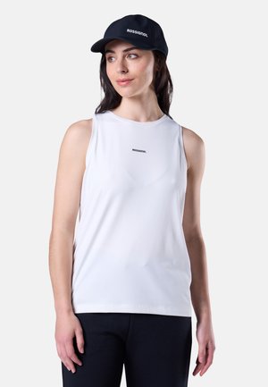 Frau trägt ein weißes ärmelloses Rossignol-Oberteil, marineblaue Hose und eine marineblaue Rossignol-Kappe, schaut leicht zur Seite.